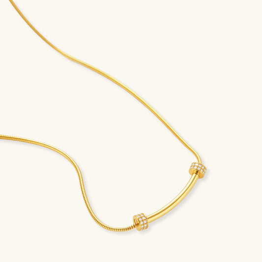 Delicate gold-plated necklace with curved bar pendant and crystal accents on snake chain against cream background.