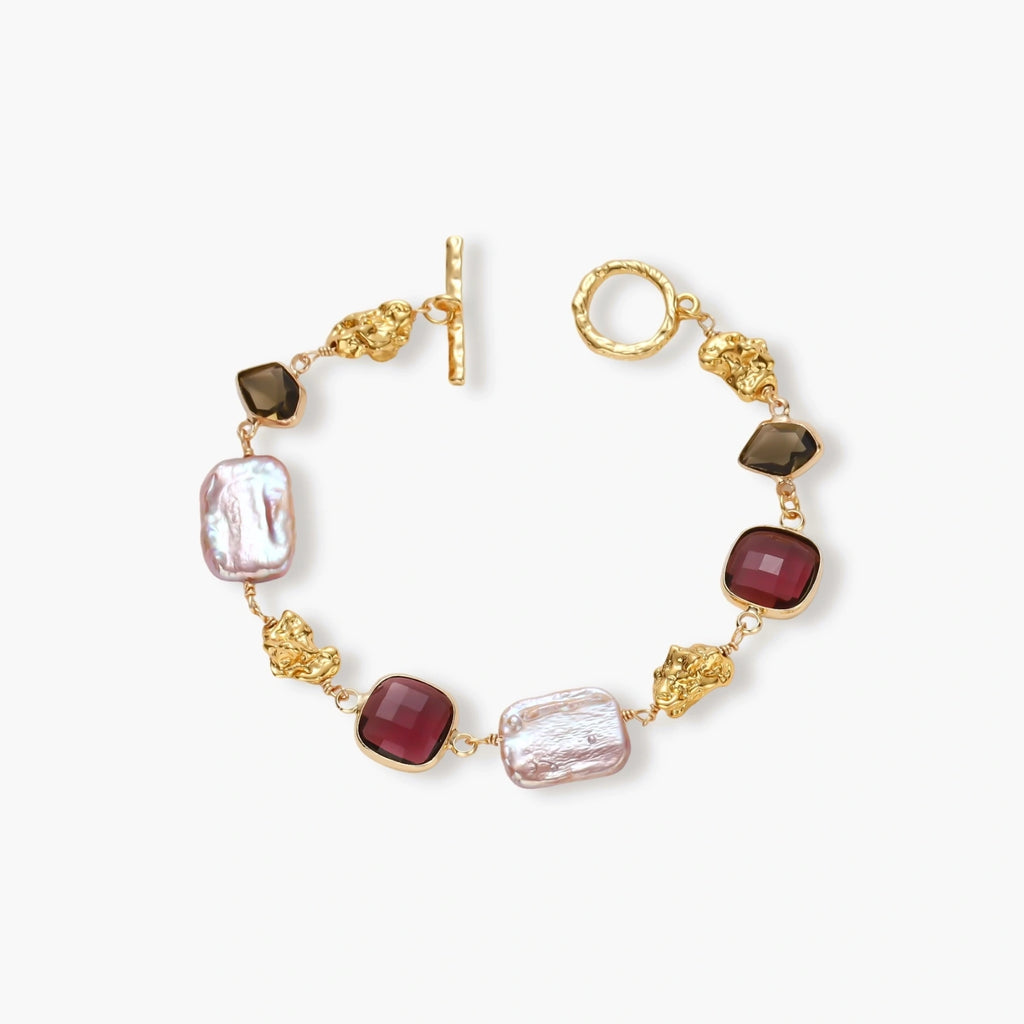 18k gold-plated copper bracelet with burgundy stones, smoky quartz, baroque pearls and ornate golden charms on white background.