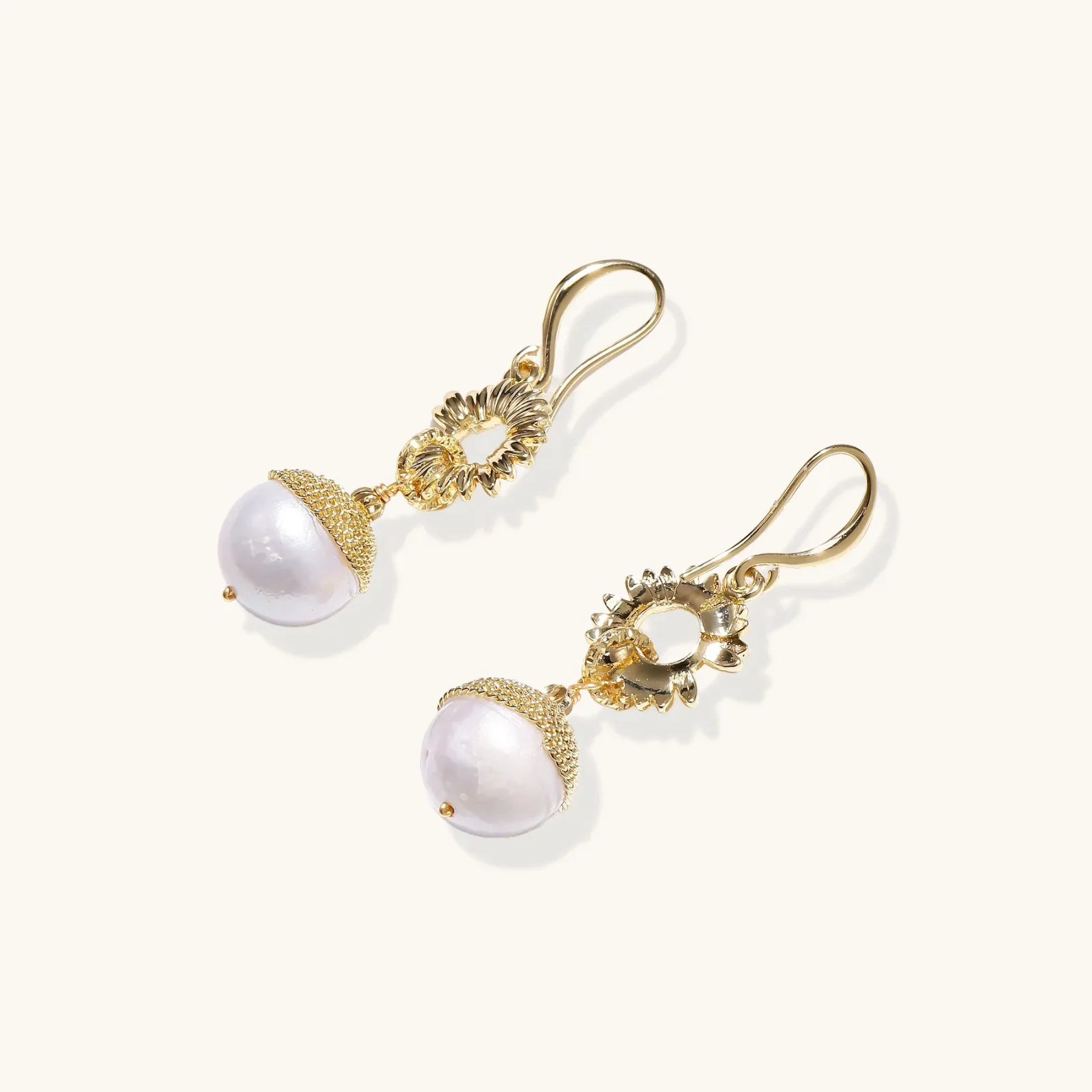Gold sunburst charm earrings with baroque pearl drops on French hooks against cream background