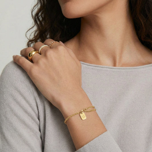 Delicate rolo-chain bracelet in gold featuring an engraved tag, elegantly worn on a wrist with additional rings.