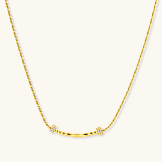 Delicate gold-plated necklace with curved bar pendant and crystal accents on snake chain against cream background.