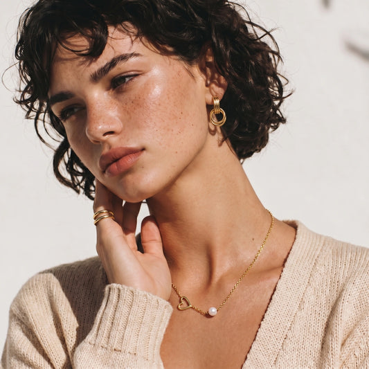 Model showcasing a slim gold necklace featuring an open heart design and a lustrous white pearl, styled with a neutral sweater.