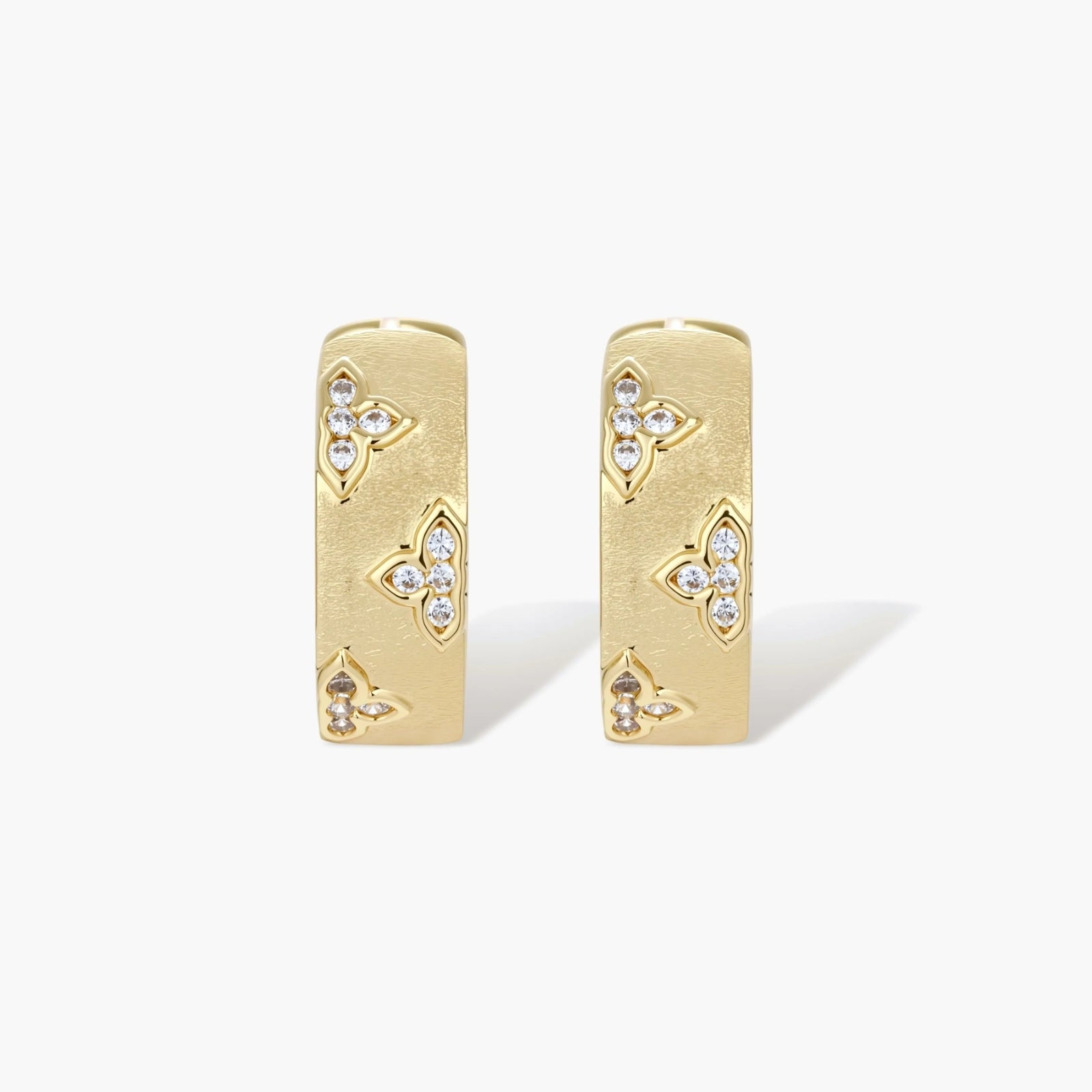 Gold-plated hoop earrings with textured surface and raised floral motifs set with clear zirconia stones on cream background.