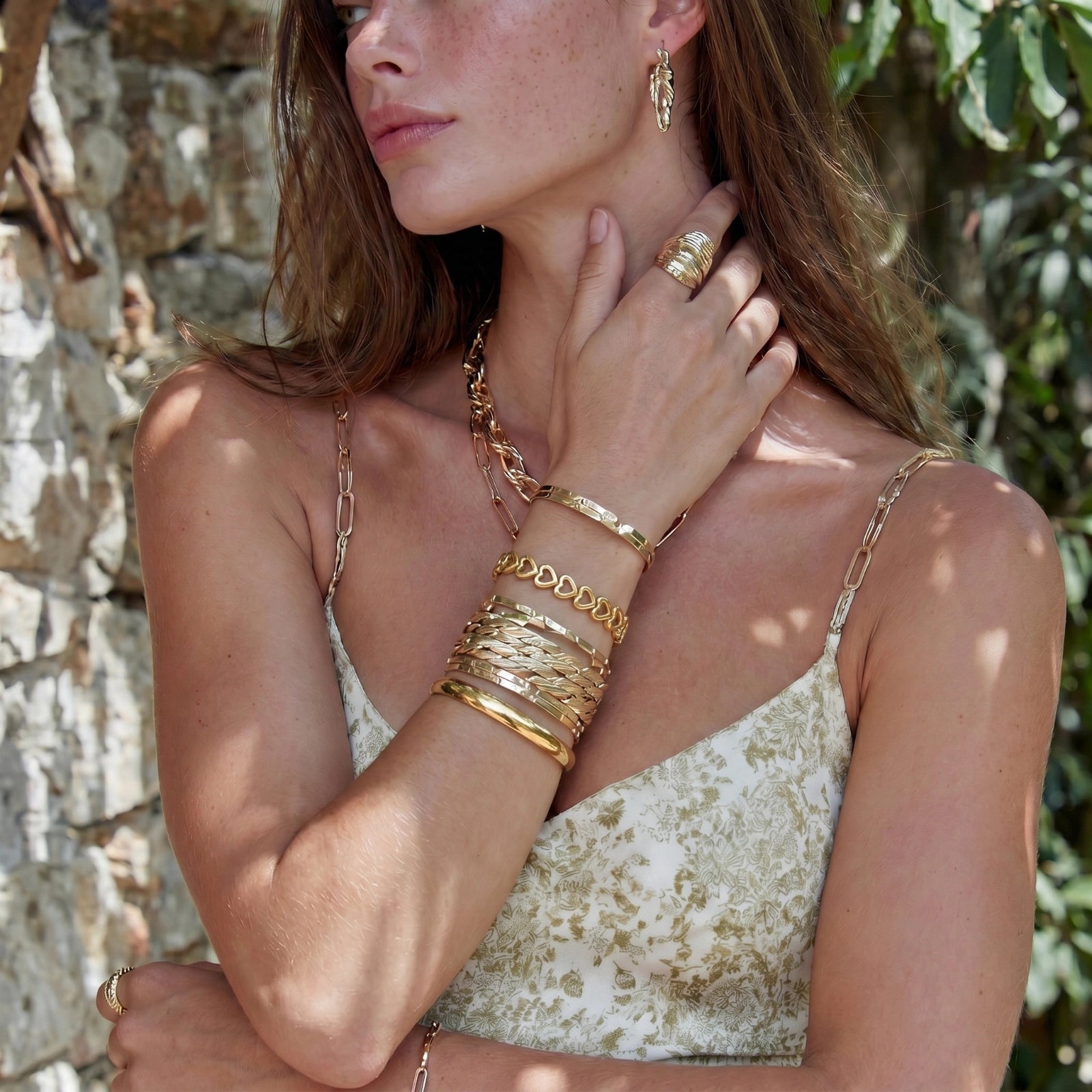 A woman showcases a radiant gold bangle featuring elegantly linked heart shapes, complementing her stylish outdoor ensemble.