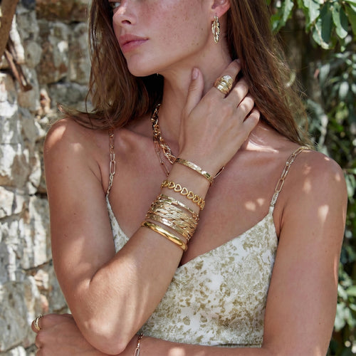 A woman showcases a radiant gold bangle featuring elegantly linked heart shapes, complementing her stylish outdoor ensemble.