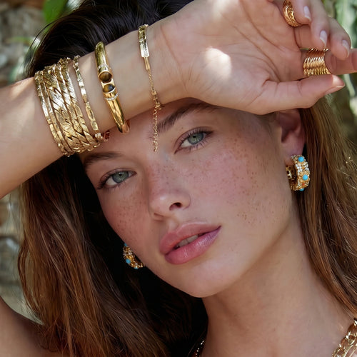 Bangle adorned with two oval tiger's eye stones, elegantly styled with gold tones and layered with other bracelets in a lush outdoor setting.