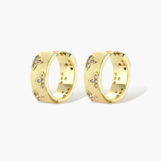 Gold-plated hoop earrings with textured surface and raised floral motifs set with clear zirconia stones on cream background.