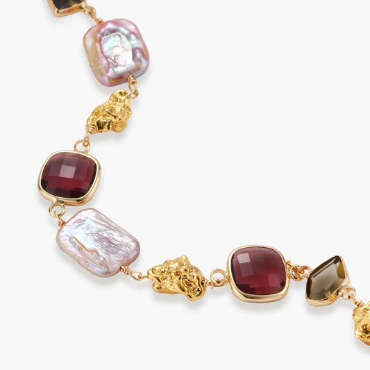 18k gold-plated copper bracelet with burgundy stones, smoky quartz, baroque pearls and ornate golden charms on white background.